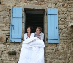 a man and woman standing in the window of an old stone building with blue shutters