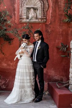 a man and woman standing next to each other in front of a red wall holding a small dog