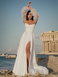 a woman in a white dress standing on rocks near the water with her arms up