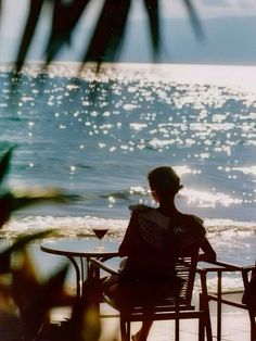 a woman sitting at a table on the beach looking out into the ocean with her back to the camera