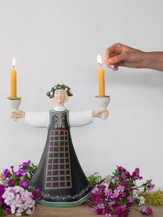 a person holding two candles in front of a figurine with flowers on the table