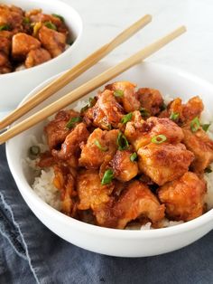 two white bowls filled with chicken and rice on top of a blue napkin next to chopsticks