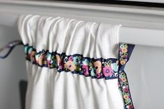 a white curtain with colorful flowers on it hanging from a rail in front of a window