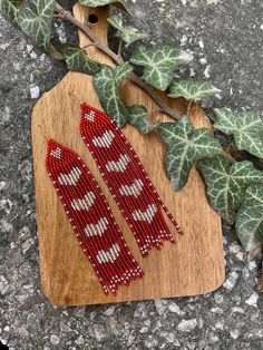 two red and white beaded earrings sitting on top of a wooden board next to ivy