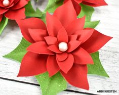 red paper flowers with green leaves and pearls on them, sitting on a white wooden surface