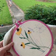 a person holding up a small embroidery project in front of some grass and bushes with yellow flowers on it