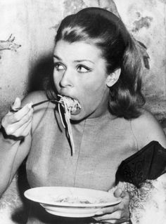 a black and white photo of a woman eating noodles with chopsticks in her mouth