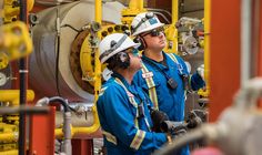 two men in hardhats and safety gear standing next to each other near pipes