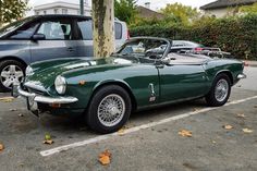 an old green sports car parked in a parking lot next to two other cars on the street