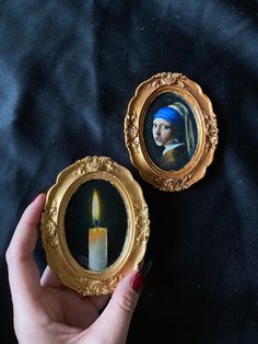 a woman holding a candle in front of two paintings on a black cloth covered surface