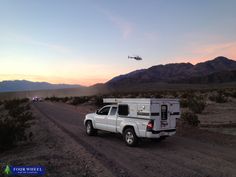 a white truck driving down a dirt road with a helicopter in the sky above it