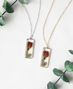 two necklaces with flowers in them on a white surface next to green plants and leaves