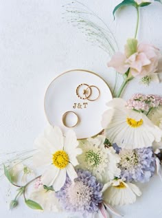 two wedding rings are placed on a plate surrounded by flowers and daisies, next to a couple's engagement ring