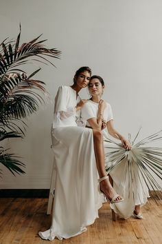 two women in white dresses are posing for a photo with palm trees behind them and one woman is holding a fan