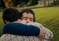 two young men hugging each other in front of a green field with houses behind them