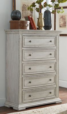 a white dresser with three vases on top of it next to a mirror and framed pictures