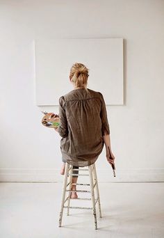 a woman sitting on a stool and holding an easel in front of a white wall