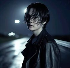 a man with black hair standing in the street at night wearing a leather jacket and looking off into the distance