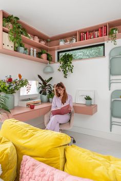 a woman sitting on a yellow couch in a room with pink furniture and plants around her