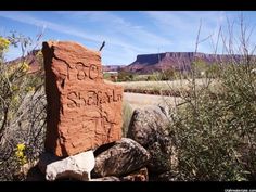 a large rock with writing on it in the middle of some bushes and mountains behind it