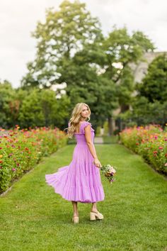 a woman in a purple dress is standing on the grass and holding a flower bouquet