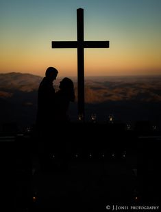 a man and woman standing in front of a cross with the sun setting behind them