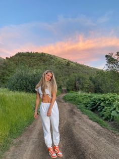 a woman standing in the middle of a dirt road with grass and bushes behind her