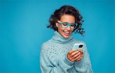 a woman wearing blue glasses looking at her cell phone while standing in front of a blue background