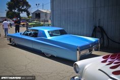 an old blue car parked in front of a building with people walking around the parking lot