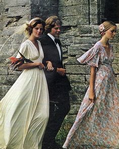 two women and a man are walking in front of a stone wall wearing long dresses