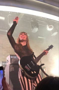 a woman in striped pants and black shirt holding up her guitar while standing on stage