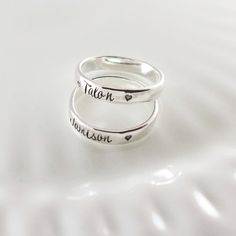 two silver rings with names on them sitting on a white surface, one has a heart in the middle