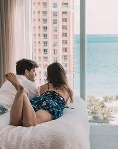 a man and woman are sitting on a bed looking out the window at the ocean