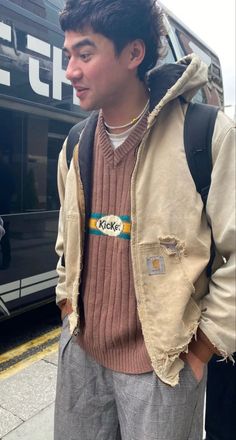 a young man standing on the sidewalk in front of a bus wearing a jacket and sweater