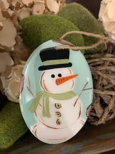 a plate with a snowman painted on it next to some flowers and moss in the background