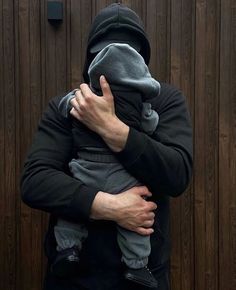 a man wearing a hoodie holding a baby in front of a wooden wall with a clock on it