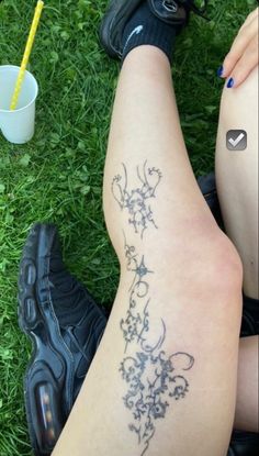 a woman's legs with tattoos on them sitting in the grass next to a drink