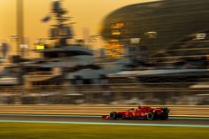 a red race car driving on a track in front of a large ship and buildings