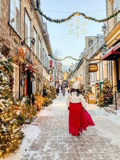 a woman walking down a snowy street with christmas decorations on the buildings and trees around her