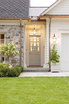 a white house with a gray door and two plants on the front lawn in front of it