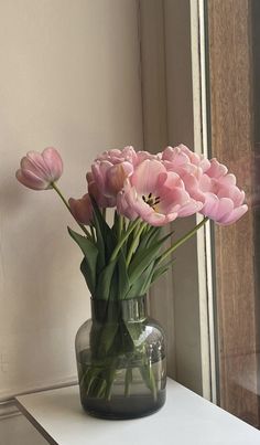 pink tulips in a glass vase on a white table next to a window