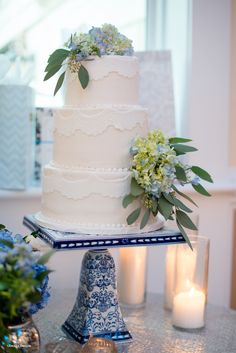 a white wedding cake with blue flowers and greenery on top sits on a table next to candles