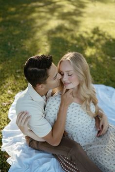 a man and woman sitting on top of a blanket in the grass kissing each other