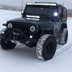 a black jeep driving through the snow in front of a car with its lights on