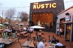 people are sitting at tables outside in front of the rustic building with lights strung over it