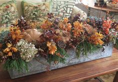 an arrangement of flowers and plants in a metal container on a table with pillows behind it