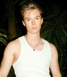 a young man wearing a white tank top standing in front of palm trees