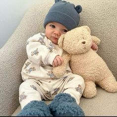 a baby sitting on a couch with a teddy bear in his lap and wearing a hat