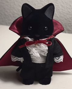 a small black cat dressed in a red cape and white lace dress, sitting on a table