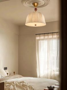 a white bed sitting under a light fixture in a bedroom next to a curtained window